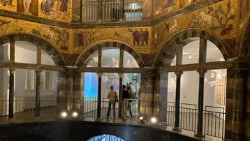 Blick in die Rotunde des Museum Wiesbaden. Die Kuppel und Wände werden geprägt durch goldenes Mosaik, das unterbrochen wird durch bunte Wappen, ebenfalls aus Mosaiksteinen. Der untere Teil des Bildes zeigt Rundbögen, durch die Blicke in die Geschosse des Museums möglich werden.