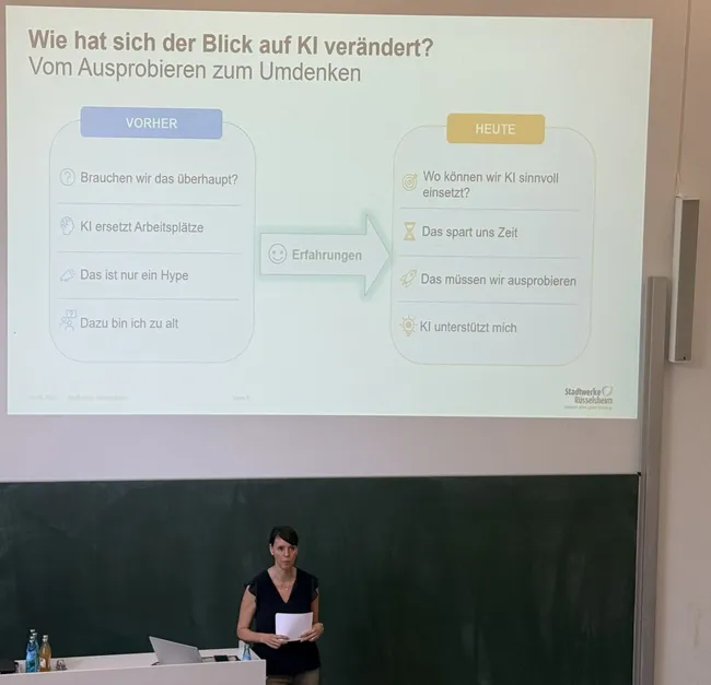 Eine Präsentation, die die Veränderung der Einstellung zu Künstlicher Intelligenz (KI) thematisiert. Links werden frühere Ansichten beschrieben, wie das Ersetzen von Arbeitsplätzen, während rechts aktuelle Perspektiven stehen, die den sinnvollen Einsatz und die Unterstützung durch KI betonen. Eine Referentin steht im Vordergrund.