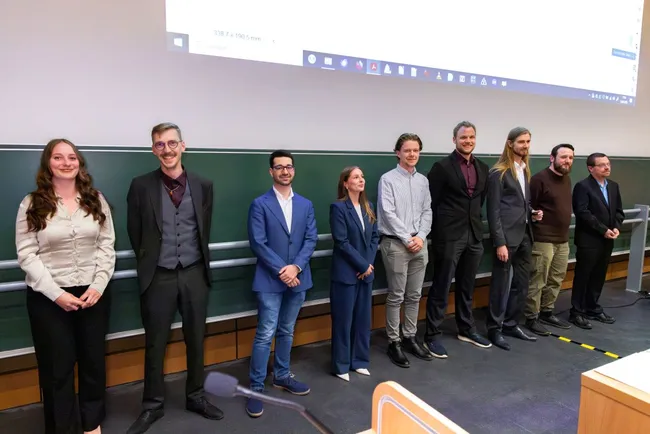 Eine Gruppe von neun Personen steht in eleganter Kleidung nebeneinander auf einer Bühne. Sie scheinen an einer Veranstaltung oder Preisverleihung teilzunehmen. Im Hintergrund ist eine Wandtafel zu sehen.