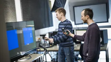 Zwei Personen in einem Raum mit Technologie arbeiten zusammen. Eine Person steht vor einem Monitor und schaut auf den Bildschirm, während die andere Person ein Gerät in der Hand hält. Im Hintergrund sind Audio-Equipment und weitere technische Geräte zu sehen.