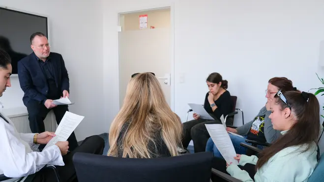 Eine Gruppe von fünf Personen sitzt in einem Raum, während ein Mann im Anzug steht und Informationen oder Anweisungen gibt. Alle Teilnehmer halten Papiere in der Hand und scheinen aufmerksam zuzuhören. Der Raum hat eine helle, moderne Atmosphäre.