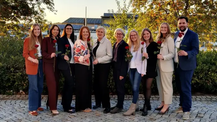 Eine Gruppe von elf Personen steht im Freien, umgeben von Bäumen. Alle halten eine Rose in der Hand und lächeln in die Kamera. Es gibt eine Vielzahl von Outfits, von casual bis elegant. Der Hintergrund zeigt einen klaren Himmel und Gebäude.