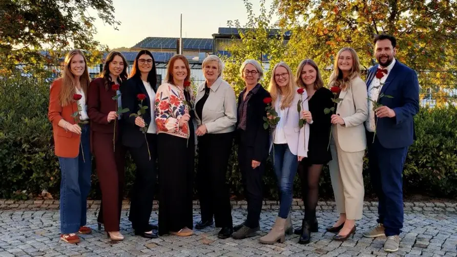Eine Gruppe von elf Personen steht im Freien, umgeben von Bäumen. Alle halten eine Rose in der Hand und lächeln in die Kamera. Es gibt eine Vielzahl von Outfits, von casual bis elegant. Der Hintergrund zeigt einen klaren Himmel und Gebäude.