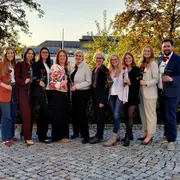 Eine Gruppe von elf Personen steht im Freien, umgeben von Bäumen. Alle halten eine Rose in der Hand und lächeln in die Kamera. Es gibt eine Vielzahl von Outfits, von casual bis elegant. Der Hintergrund zeigt einen klaren Himmel und Gebäude.