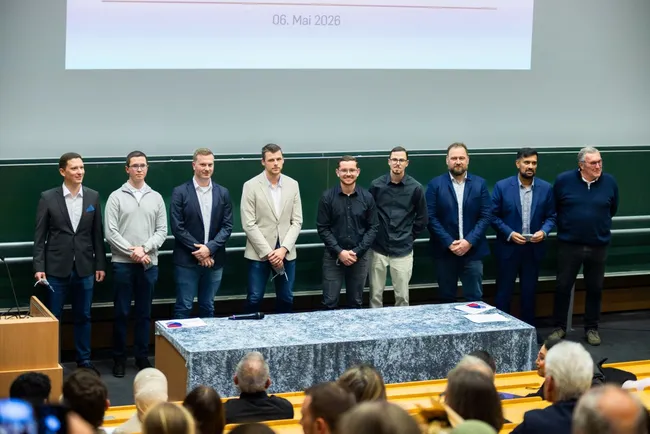 Eine Gruppe von neun Männern steht auf einer Bühne vor einem Publikum in einem Konferenzsaal. Sie tragen verschiedene formelle Kleidung. Auf dem Tisch vor ihnen liegt ein Tuch. Im Hintergrund ist ein Präsentationsbildschirm mit dem Datum „06. Mai 2026“ zu sehen.