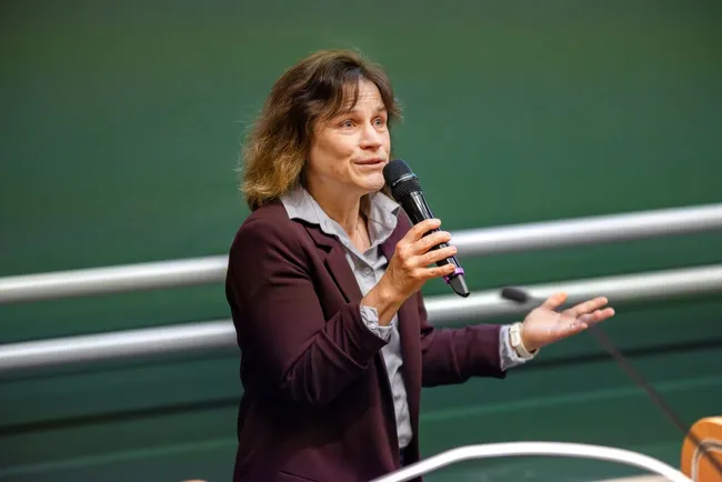 Eine Frau mit lockigem Haar spricht in ein Mikrofon, während sie gestikuliert. Sie trägt ein dunkles Sakko über einem hellen Hemd. Der Hintergrund ist unscharf, aber scheint eine öffentliche Veranstaltung zu sein.