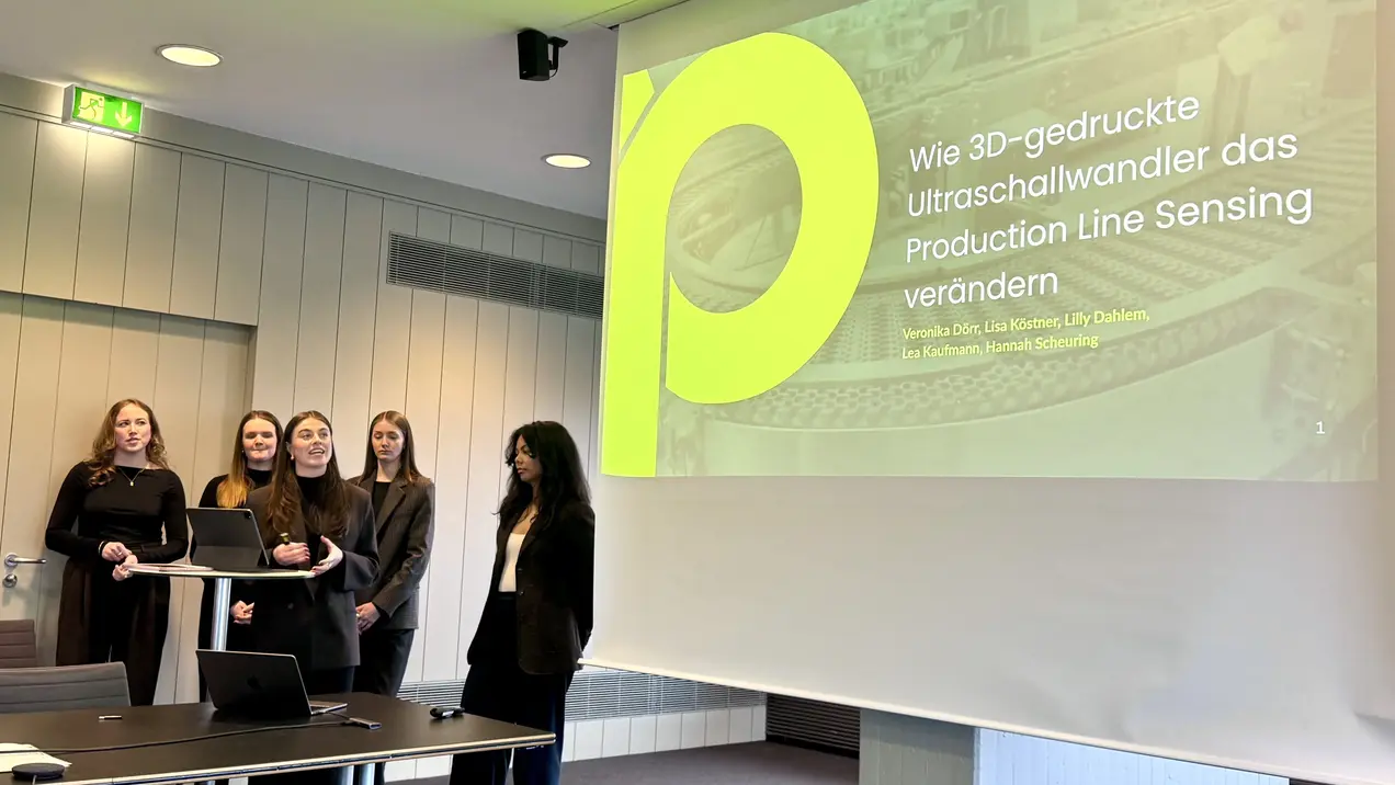 Fünf Frauen stehen in formeller Kleidung vor einem Präsentationstisch, während auf einer Leinwand der Titel "Wie 3D-gedruckte Ultraschallwandler das Production Line Sensing verändern" zu sehen ist. Die Szene vermittelt eine professionelle Atmosphäre einer Präsentation oder Konferenz.