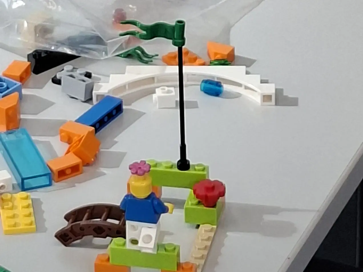 Auf dem Tisch steht eine LEGO-Baufigur mit einer blauen Kleidung, einem rosa Hut und einer Blume. Sie ist auf einer kleinen, bunten Plattform platziert, die von grünen und orangefarbenen LEGO-Steinen umgeben ist. Hinter ihr weht eine kleine grüne Flagge.