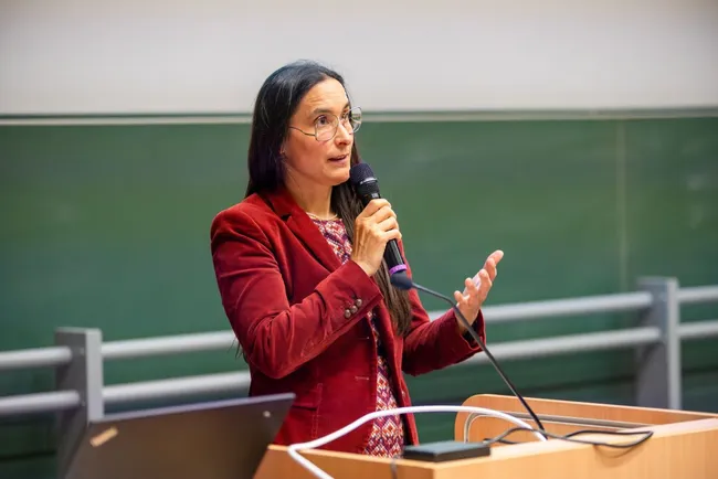 Eine Frau mit langen, dunklen Haaren und einer roten Blazerjacke steht am Mikrofon und spricht. Hintergrund ist eine grüne Wandtafel in einem Seminarraum. Sie gestikuliert, während sie ihre Punkte verdeutlicht.