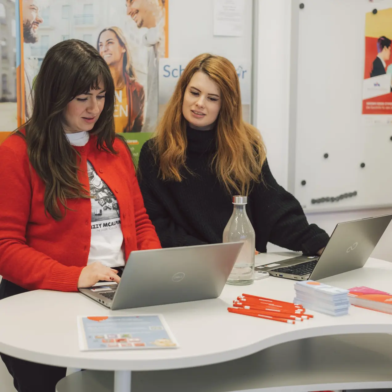 Zwei Frauen sitzen an einem Tisch und schauen auf ihre Laptops. Eine Frau trägt einen roten Pullover, die andere einen schwarzen Pullover. Auf dem Tisch liegen Broschüren und ein Wasserkrug. Im Hintergrund sind Informationsplakate sichtbar.