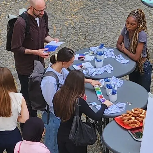 Eine Gruppe von Menschen steht um einen Tisch, auf dem Informationsmaterialien und einige Gegenstände ausgelegt sind. Einige Personen sprechen miteinander, während andere sich Notizen machen. Im Hintergrund ist ein Werbebanner zu sehen, das eine Institution bewirbt.