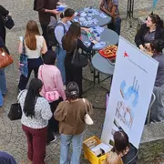 Eine Gruppe von Menschen steht um einen Tisch, auf dem Informationsmaterialien und einige Gegenstände ausgelegt sind. Einige Personen sprechen miteinander, während andere sich Notizen machen. Im Hintergrund ist ein Werbebanner zu sehen, das eine Institution bewirbt.