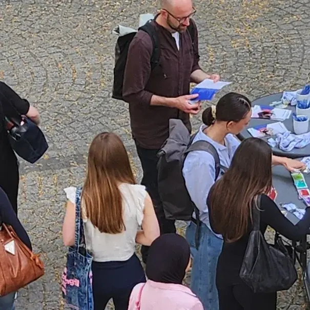 Eine Gruppe von Menschen steht um einen Tisch, auf dem Informationsmaterialien und einige Gegenstände ausgelegt sind. Einige Personen sprechen miteinander, während andere sich Notizen machen. Im Hintergrund ist ein Werbebanner zu sehen, das eine Institution bewirbt.