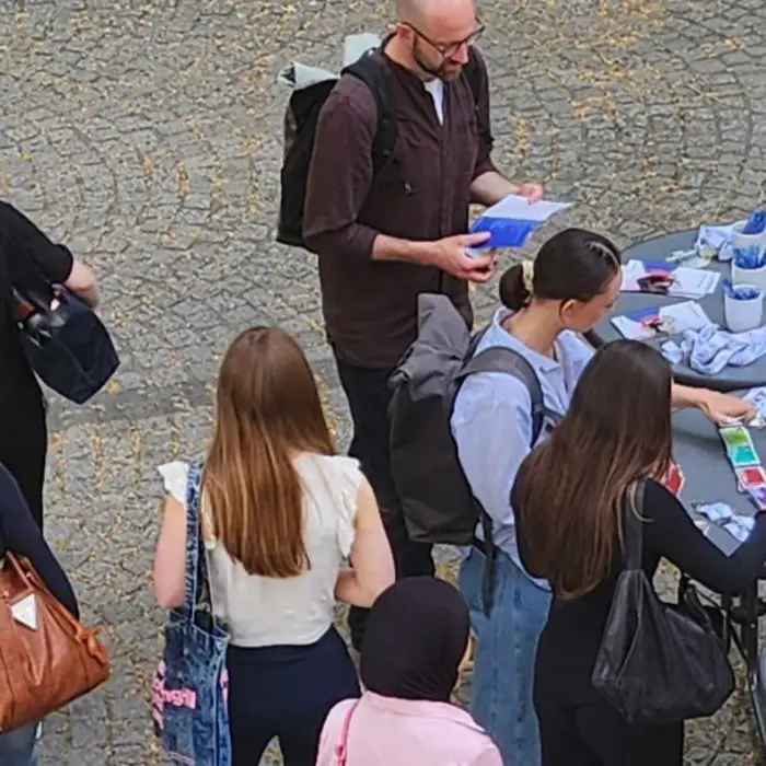 Eine Gruppe von Menschen steht um einen Tisch, auf dem Informationsmaterialien und einige Gegenstände ausgelegt sind. Einige Personen sprechen miteinander, während andere sich Notizen machen. Im Hintergrund ist ein Werbebanner zu sehen, das eine Institution bewirbt.