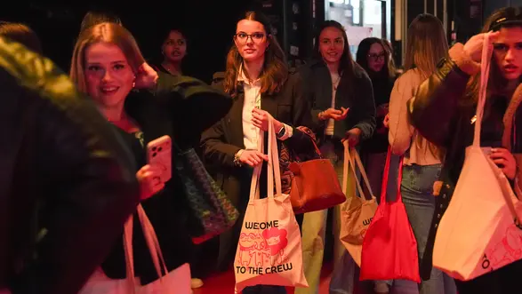 Gruppe junger Frauen steht an einer Eingangstür in einem belebten Raum. Einige halten Stofftaschen und Smartphones. Sie lächeln und wirken gespannt, während sie auf den Eintritt warten. Die Beleuchtung ist warm und einladend.