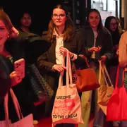 Gruppe junger Frauen steht an einer Eingangstür in einem belebten Raum. Einige halten Stofftaschen und Smartphones. Sie lächeln und wirken gespannt, während sie auf den Eintritt warten. Die Beleuchtung ist warm und einladend.