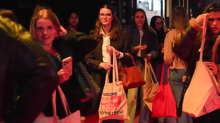 Gruppe junger Frauen steht an einer Eingangstür in einem belebten Raum. Einige halten Stofftaschen und Smartphones. Sie lächeln und wirken gespannt, während sie auf den Eintritt warten. Die Beleuchtung ist warm und einladend.
