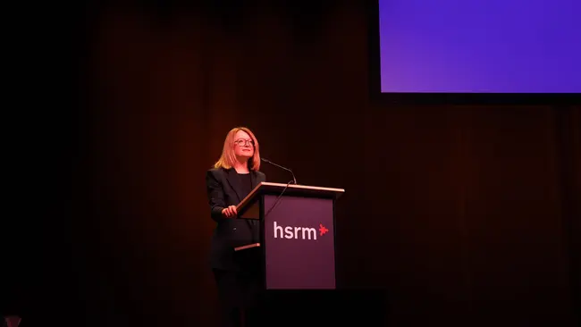 Eine Frau steht an einem Podium mit der Aufschrift "hsrm". Sie lächelt und spricht zu einem Publikum. Im Hintergrund ist ein großer Bildschirm zu sehen, der leer ist. Die Atmosphäre wirkt professionell und einladend.