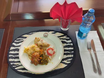 Auf dem Tisch liegt ein Teller mit einem vielfältigen Gericht: gebratenes Hühnchen, Reis, Gemüse und Nudeln. Daneben steht eine Flasche Wasser und ein Glas mit einer roten Serviette. Der Teller befindet sich auf einem runden, schwarz-weißen Platzset.