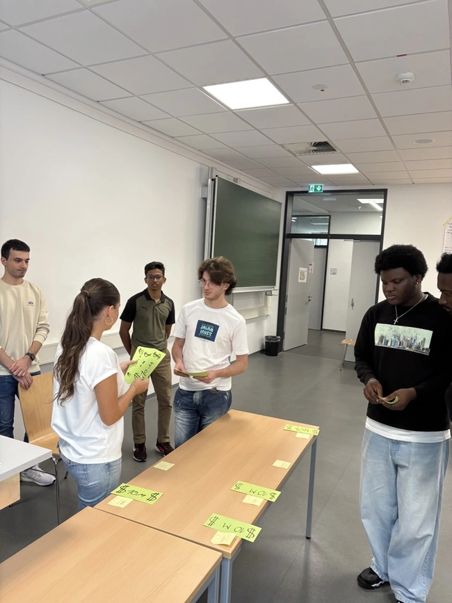 Eine Gruppe von sechs Schülern steht um zwei Tische in einem Klassenzimmer. Auf den Tischen liegen grüne Karten mit Zahlen. Ein Mädchen in einem weißen T-Shirt spricht mit einem Jungen, während die anderen aufmerksam zuhören.