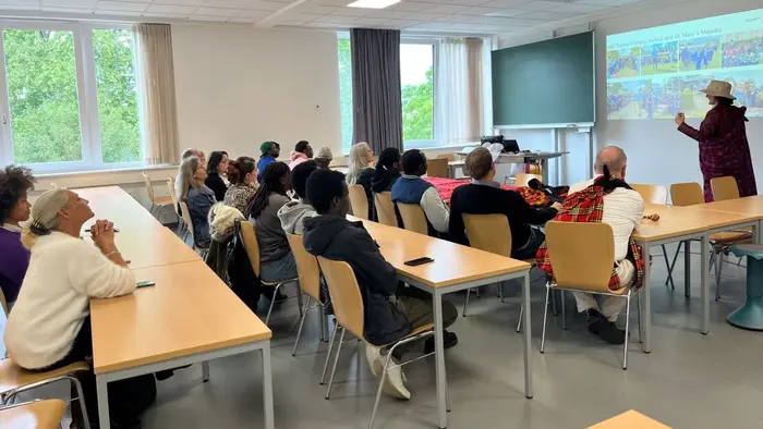 Eine Gruppe von Menschen sitzt an Tischen in einem Klassenraum und hört einem Redner zu, der an eine Leinwand mit einer Präsentation zeigt. Fenster im Hintergrund lassen Tageslicht herein. Die Atmosphäre ist konzentriert und informativ.