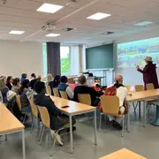 Eine Gruppe von Menschen sitzt an Tischen in einem Klassenraum und hört einem Redner zu, der an eine Leinwand mit einer Präsentation zeigt. Fenster im Hintergrund lassen Tageslicht herein. Die Atmosphäre ist konzentriert und informativ.