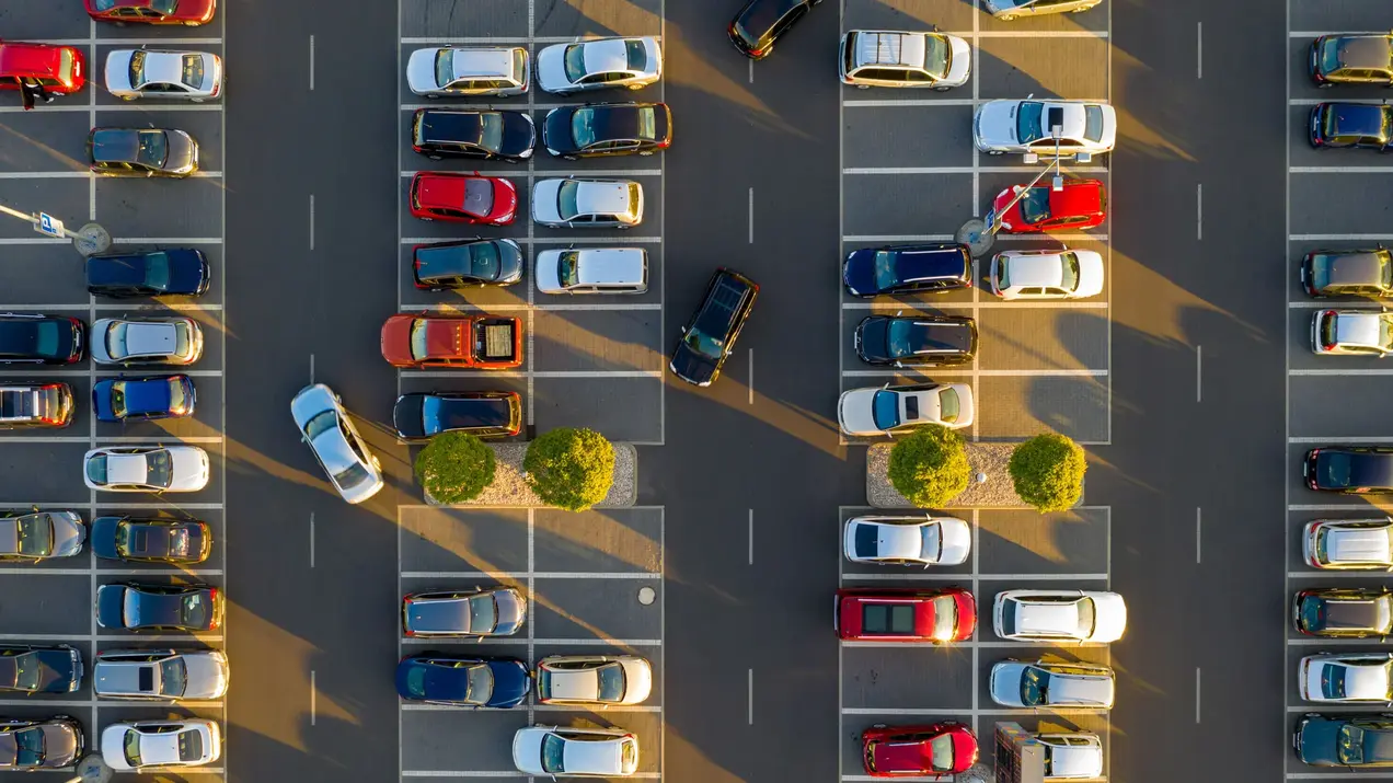 Eine Luftaufnahme eines Parkplatzes mit vielen geparkten Autos in verschiedenen Farben. Zwischen den Parklücken sind kleine Bäume zu sehen. Der Parkbereich ist übersichtlich angeordnet, mit klaren Fahrzeugreihen und einigermaßen freien Lücken.