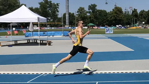 Ein junger Athlet in einem gelben Trikot läuft auf einer blauen Laufbahn. Er trägt kurze Hosen und zeigt eine dynamische Laufhaltung. Im Hintergrund ist eine Sportveranstaltung mit Zuschauern und einer Überdachung zu sehen. 