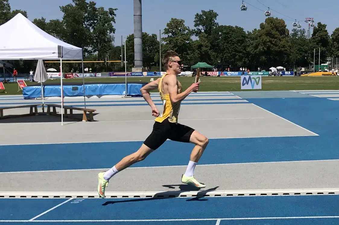 Ein junger Athlet in einem gelben Trikot läuft auf einer blauen Laufbahn. Er trägt kurze Hosen und zeigt eine dynamische Laufhaltung. Im Hintergrund ist eine Sportveranstaltung mit Zuschauern und einer Überdachung zu sehen. 