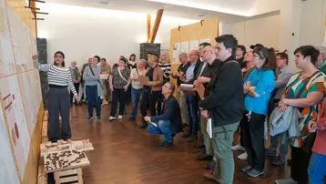 Eine Architekturstudentin steht vor einer Wand mit Postern zu ihrem Entwurf der Dorfmitte in Wallroth. Sie erklärt ihren Entwurf einer Gruppe von Menschen, die aufmerksam zuhören.