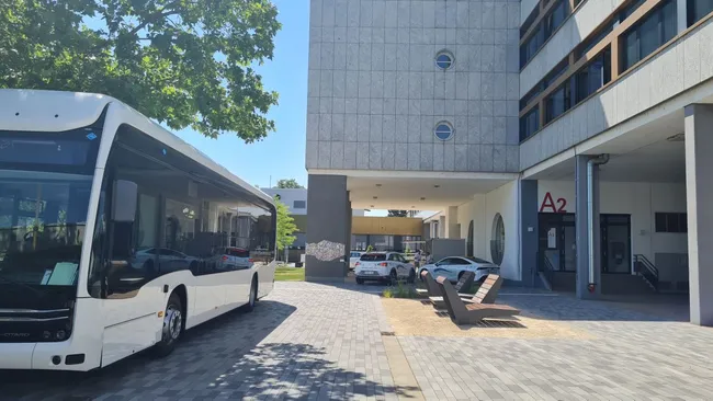 Ein weißer Bus steht auf einem gepflasterten Platz vor einem modernen Gebäude mit großen Fenstern. Rechts ist der Eingang mit der Kennzeichnung „A2“ sichtbar. Im Hintergrund sind weitere Fahrzeuge und Grünflächen zu erkennen.