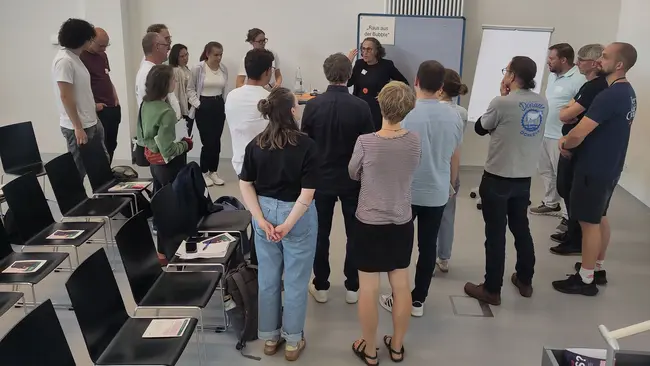 Eine Gruppe von etwa 15 Personen versammelt sich in einem weißen Raum. Eine Person steht in der Mitte und spricht, während die anderen aufmerksam zuhören. Einige Stühle sind im Hintergrund sichtbar, aber die Menschen stehen nah zusammen und sind in ein Gespräch vertieft.