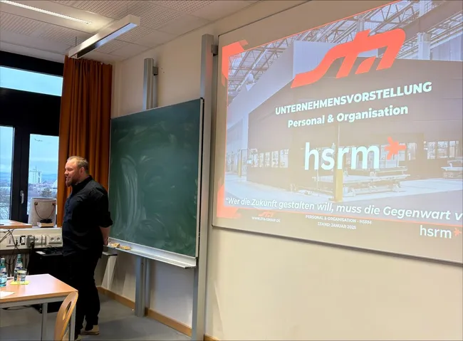 Der Bildschirm zeigt eine Unternehmensvorstellung der sta Schalltechnische Anlagen GmbH. Ein Referent steht vor der Präsentation in einem Raum mit Fenstern, die Tageslicht hereinlassen. Auf der Folie steht: „Wer die Zukunft gestalten will, muss die Gegenwart verstehen.“