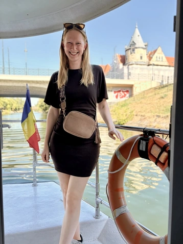 Eine lächelnde Frau mit langen, blonden Haaren steht auf einem Boot in der Nähe eines Flusses. Sie trägt ein schwarzes Kleid und hält eine kleine Tasche. Im Hintergrund sind ein Brückenbogen und ein historisches Gebäude sichtbar.