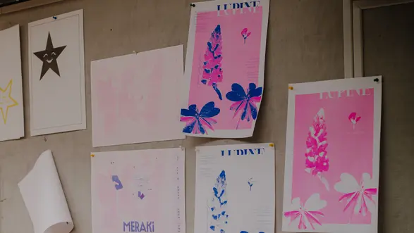 An einer Wand hängen verschiedene Kunstwerke. Die Bilder zeigen einerseits in sanften Pink- und Blautönen gestaltete Motive mit Floral-Elementen und Schriftzügen. Einige unbedruckte Blätter sind ebenfalls darunter angeordnet.