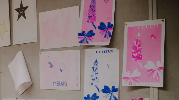 An einer Wand hängen verschiedene Kunstwerke. Die Bilder zeigen einerseits in sanften Pink- und Blautönen gestaltete Motive mit Floral-Elementen und Schriftzügen. Einige unbedruckte Blätter sind ebenfalls darunter angeordnet.