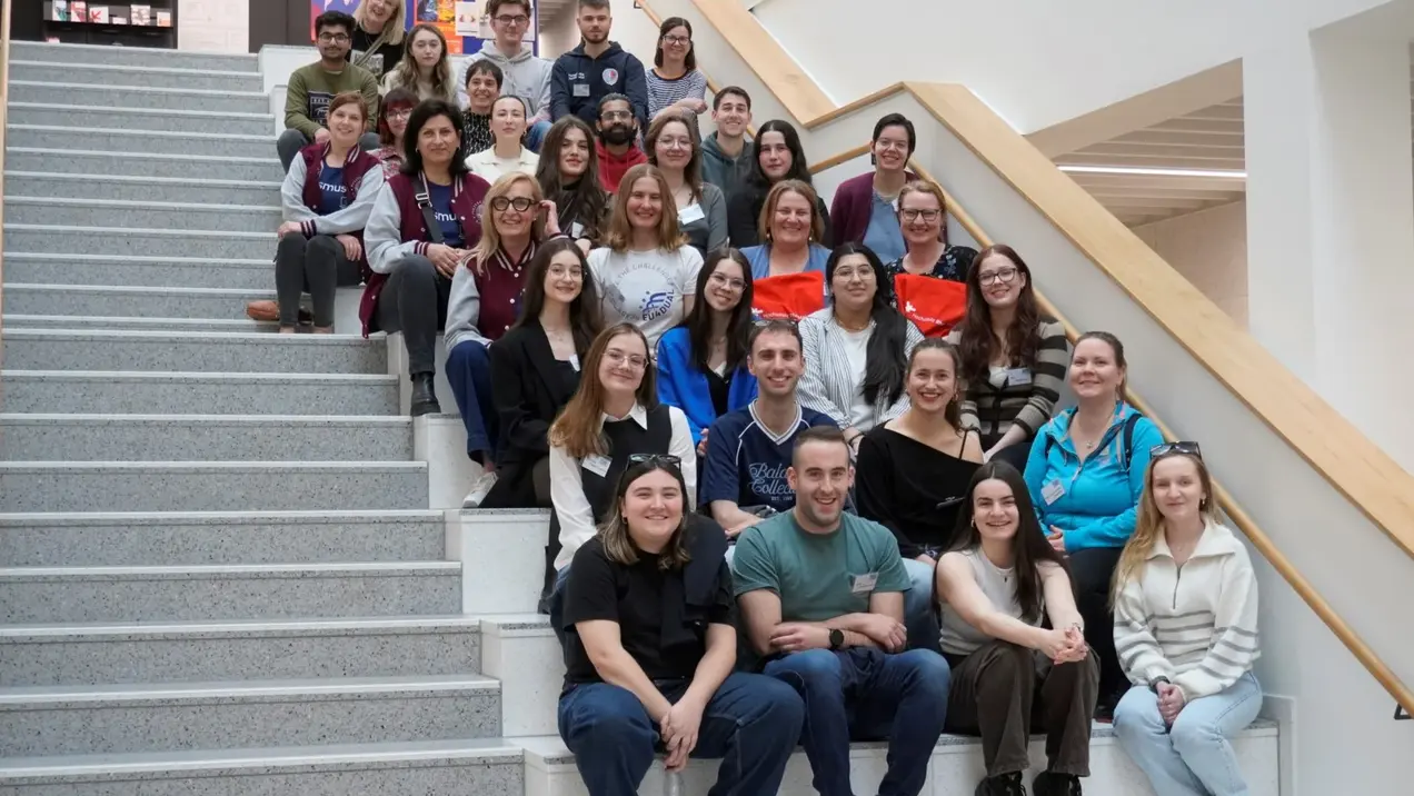 Eine Gruppe von etwa 40 Menschen sitzt und steht auf einer Treppe. Sie lächeln und schauen in die Kamera. Die Atmosphäre ist freundlich und einladend. Im Hintergrund sind Bücherregale und weitere Treppenstufen sichtbar.