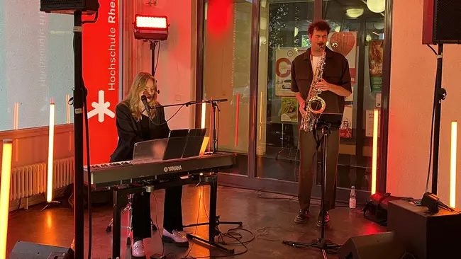 Eine Sängerin sitzt an einem Klavier, während ein Saxophonist neben ihr spielt. 