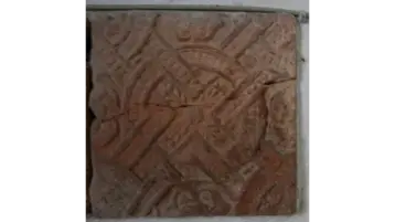 The image shows a square, decorated tile with a complex pattern. The surface texture consists of engraved geometric shapes and lines that form an artistic design. The colour nuances vary between reddish and brown tones.