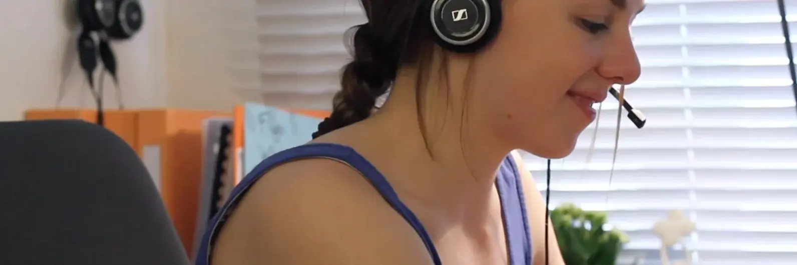 Eine junge Frau sitzt in einem Büro und trägt ein Headset. Sie lächelt leicht, während sie an einem Computer arbeitet. Im Hintergrund sind Bücher und eine Pflanze zu sehen, die eine angenehme Arbeitsumgebung schaffen.