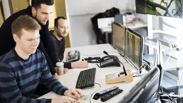 Drei Männer arbeiten gemeinsam an einem Schreibtisch mit mehreren Computermonitoren. Einer der Männer tippt auf einer farbigen Tastatur, während die anderen beiden ihn aufmerksam beobachten. Getränke und Büromaterial sind ebenfalls auf dem Tisch platziert.