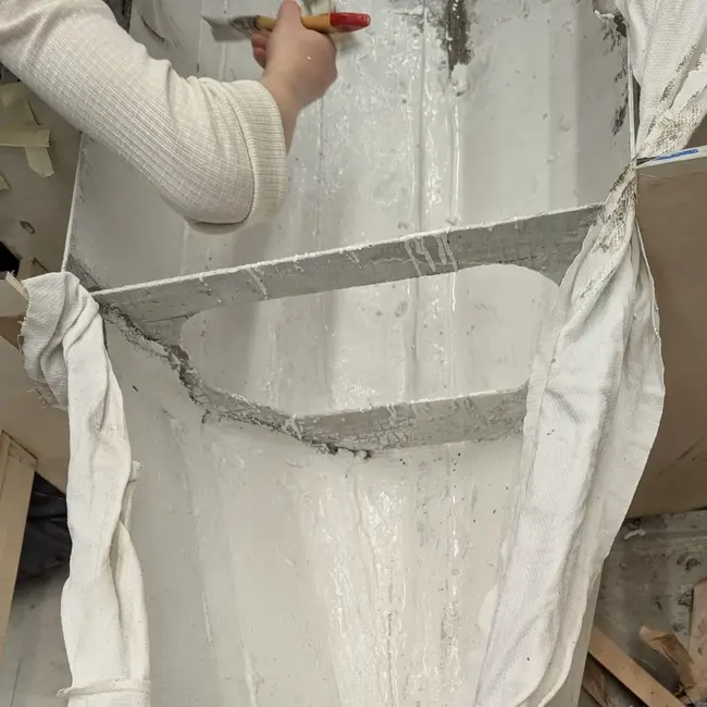 Eine Person trägt mit einem Werkzeug eine weiße, pastöse Substanz auf die Innenseite einer langen, schmalen Form auf. Die Form ist mit einem Tuch ausgekleidet und umgeben von Holzresten und Arbeitsmaterialien.