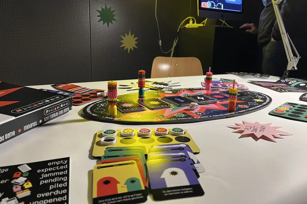 Auf dem Tisch liegt ein Brettspiel mit einem bunten Spielboard, Markern und Spielkarten. Einige Sticker mit verschiedenen Farben und Symbolen sind ebenfalls sichtbar. Im Hintergrund ist ein Bildschirm mit grafischen Elementen zu sehen, der Teil des Spieles ist.