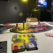 Auf dem Tisch liegt ein Brettspiel mit einem bunten Spielboard, Markern und Spielkarten. Einige Sticker mit verschiedenen Farben und Symbolen sind ebenfalls sichtbar. Im Hintergrund ist ein Bildschirm mit grafischen Elementen zu sehen, der Teil des Spieles ist.