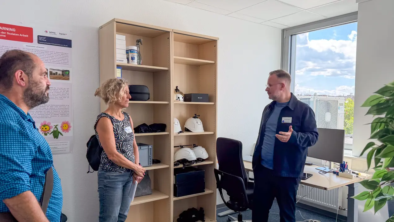 Ein Mann in Arbeitskleidung erklärt etwas, während eine Frau und ein Mann ihm zuhören. Im Hintergrund steht ein Regal mit verschiedenen Gegenständen, darunter Helme und Ausrüstung. Der Raum ist hell und freundlich, mit einem Fenster, das Tageslicht hineinlässt.