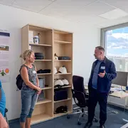 Ein Mann in Arbeitskleidung erklärt etwas, während eine Frau und ein Mann ihm zuhören. Im Hintergrund steht ein Regal mit verschiedenen Gegenständen, darunter Helme und Ausrüstung. Der Raum ist hell und freundlich, mit einem Fenster, das Tageslicht hineinlässt.