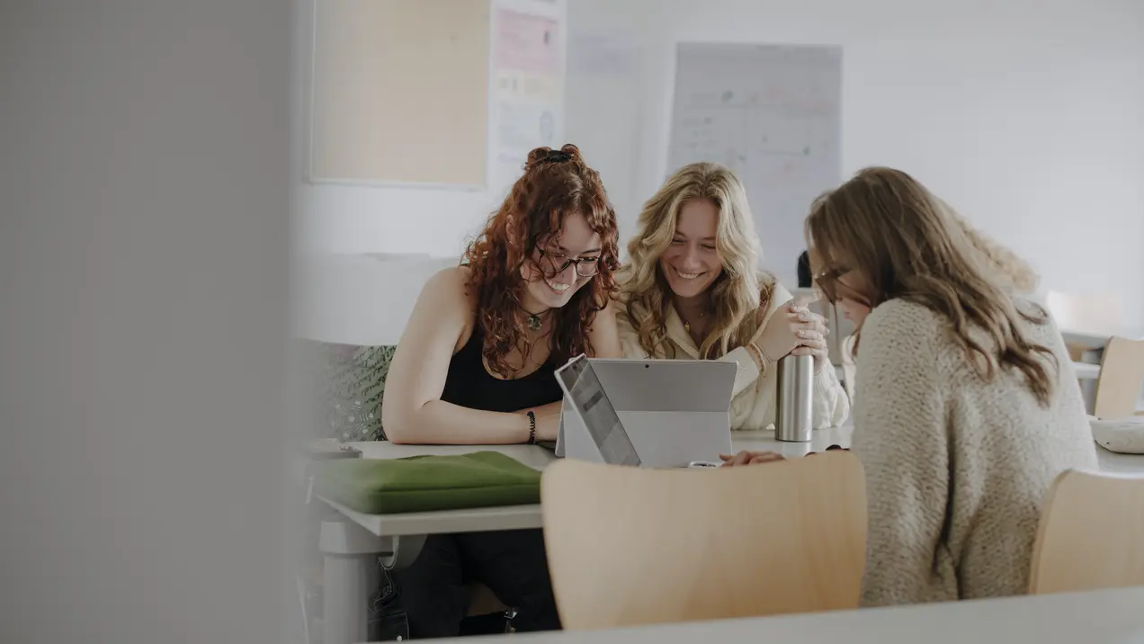 Man sieht drei Studierende im Vordergrund, die gemeinsam an einem Projekt arbeiten und lächeln. Im Hintergrund sieht man Plakate.