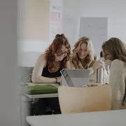 Man sieht drei Studierende im Vordergrund, die gemeinsam an einem Projekt arbeiten und lächeln. Im Hintergrund sieht man Plakate.