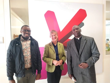 Drei Personen stehen vor einem großen, bunten Hintergrund. Links steht ein Mann mit Brille und einem gemusterten Hemd. In der Mitte steht eine Frau mit Brille und einem blauen Oberteil, rechts ein Mann in einem grauen Mantel. Alle lächeln.