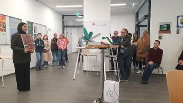 Eine Person in formeller Kleidung hält ein Kunstwerk in der Hand und spricht vor einer Gruppe von Zuschauern, die aufmerksam zuhören. Die Szene ist in einem hellen Raum mit Tischen und Stühlen, sowie Informationsplakaten an den Wänden.